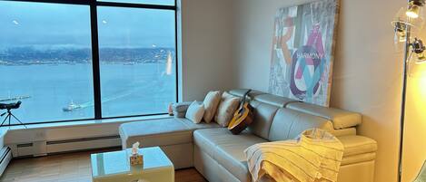 A modern living room with a large window offering a city view, a sofa with cushions, a guitar, a small table with a tissue box, and a floor lamp.