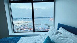 A bedroom with a large window overlooking a port with shipping containers and a distant building.