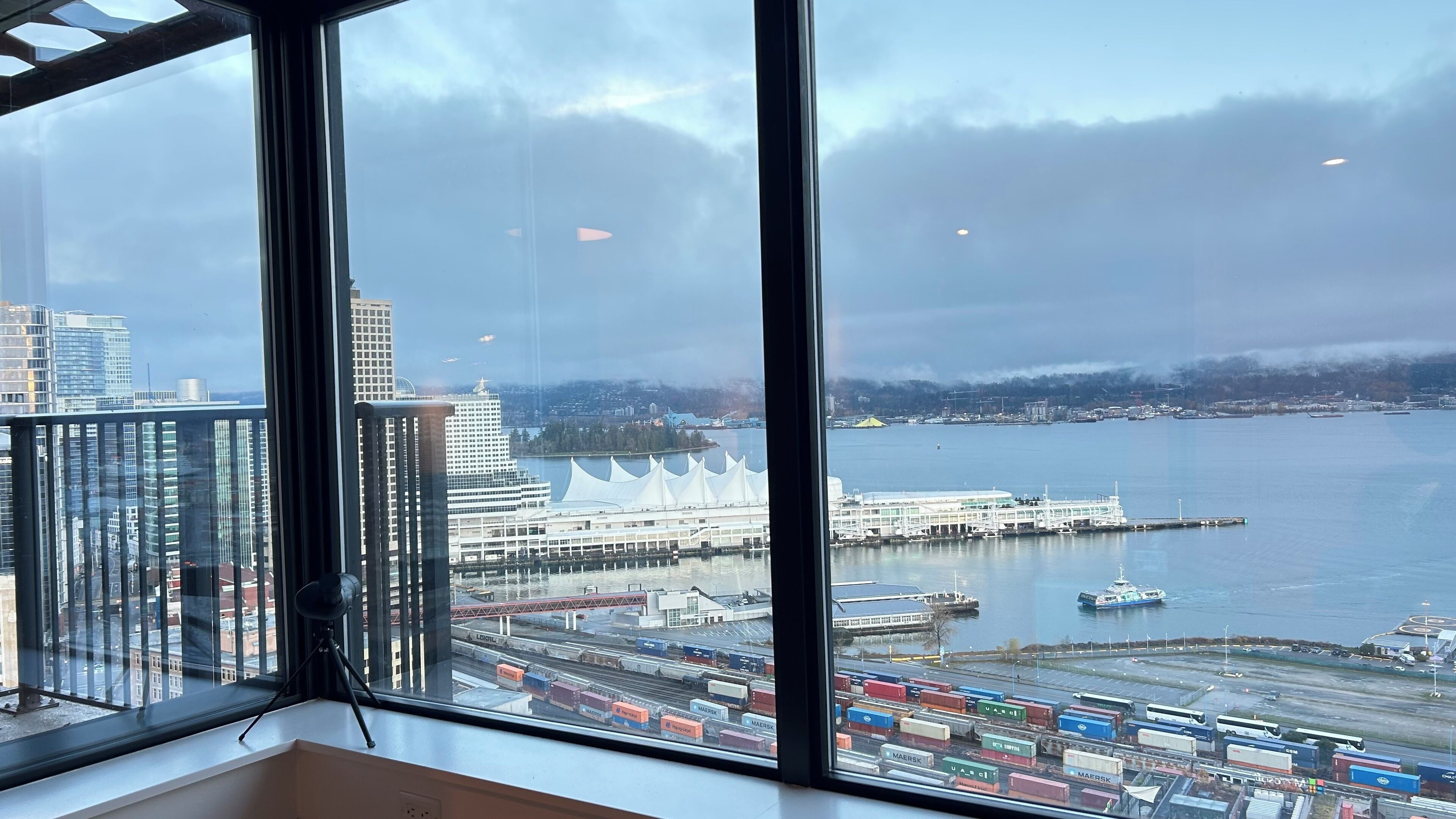 Vista desde un piso alto con vista al puerto, contenedores de carga, un crucero y un paisaje urbano de fondo.