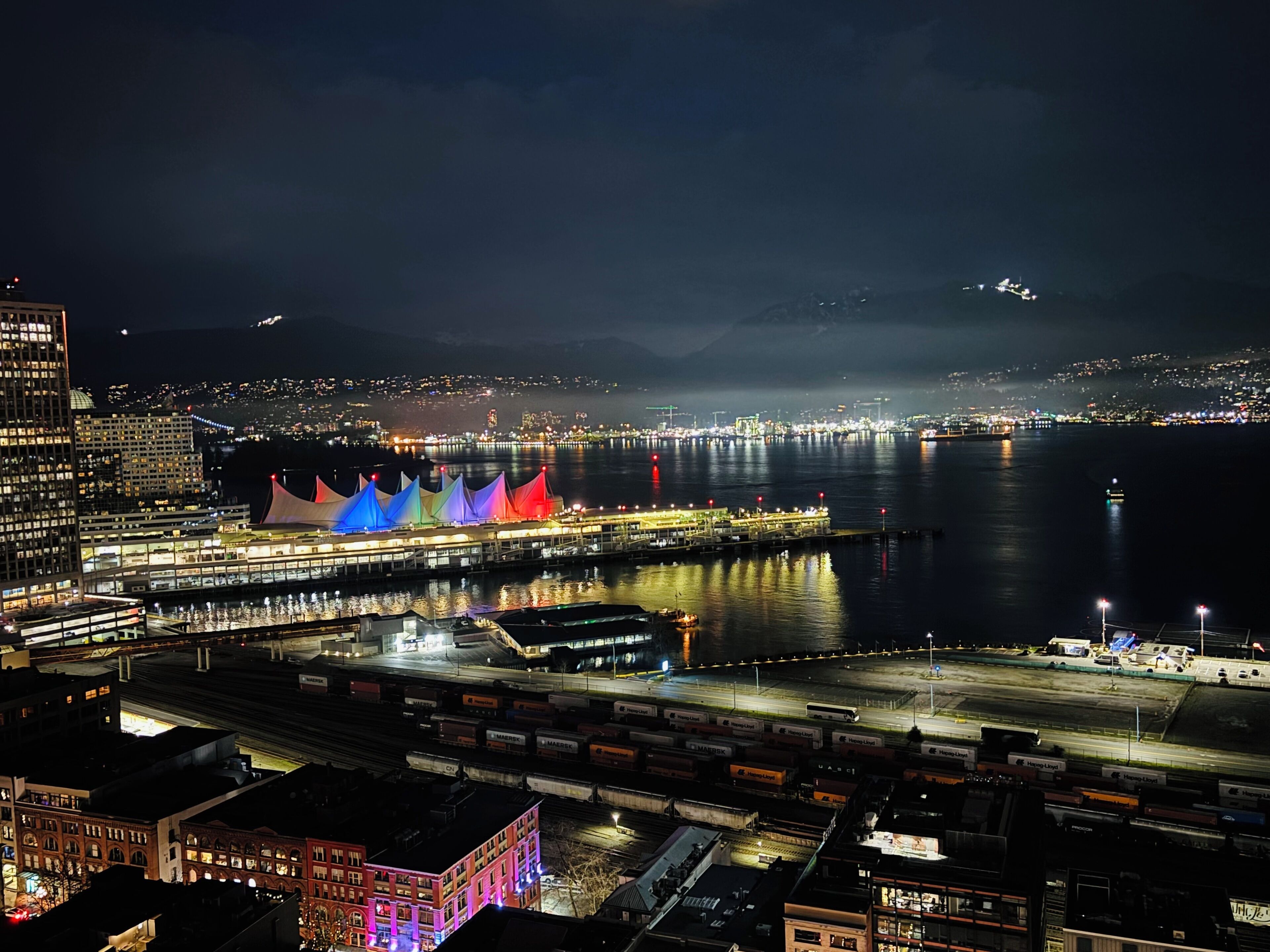 A nighttime cityscape with a waterfront promenade, illuminated buildings, and a bridge.
