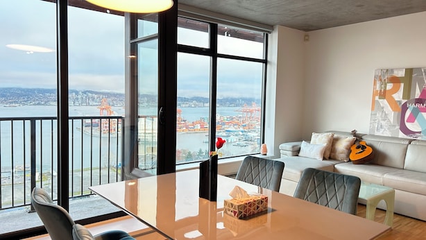 A modern living room with a large window offering a city view, a dining table, and a sofa with a guitar.