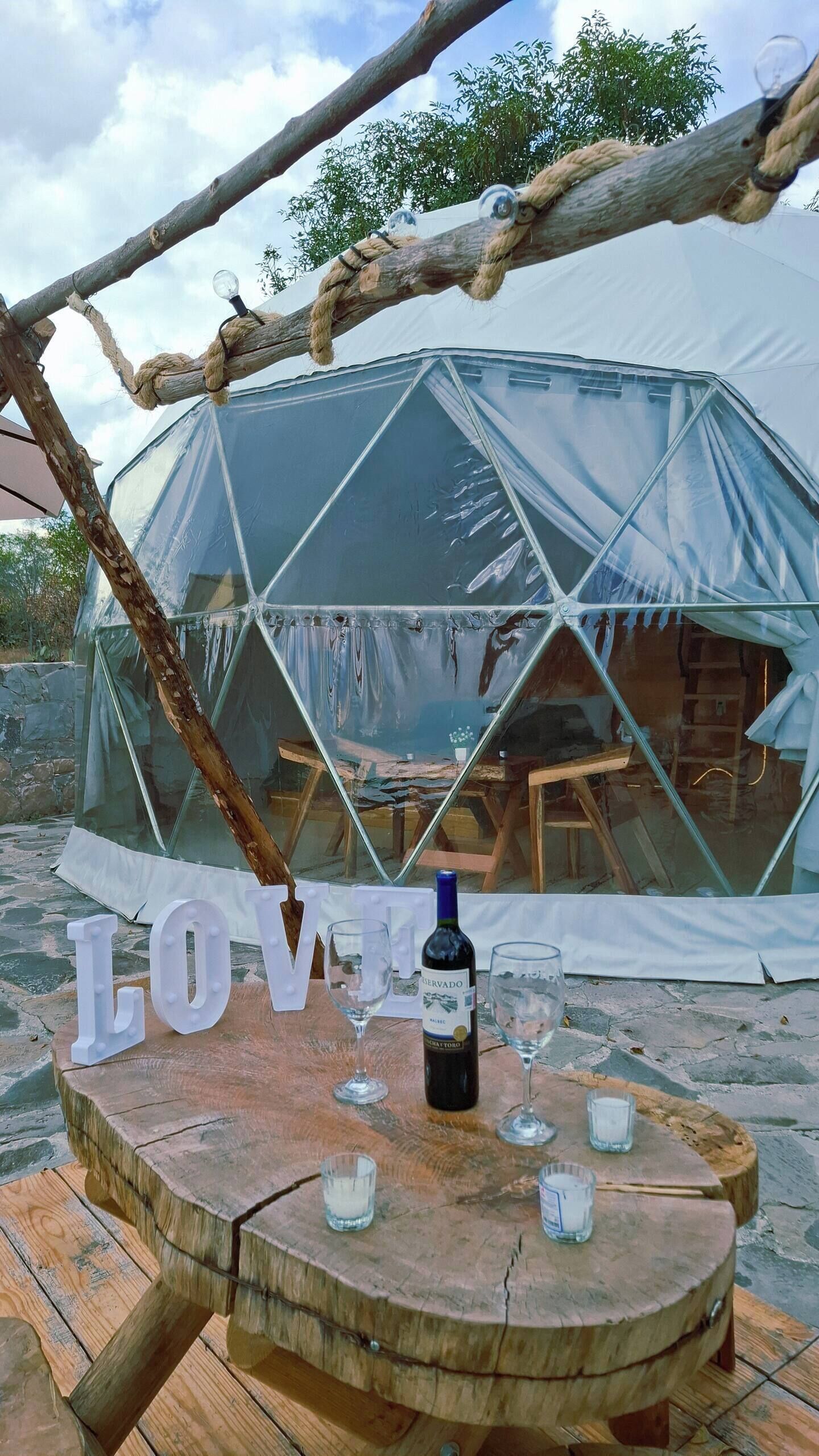 Una tienda en forma de cúpula geodésica transparente con una mesa del frente, una botella de vino y copas.