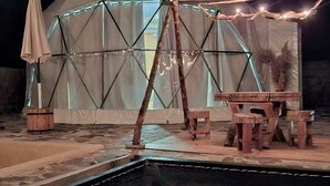 Una estructura en forma de cúpula geodèsica amb una piscina al davant, seients de fusta i llums penjades.
