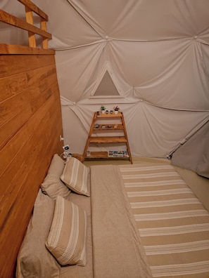 Uma tenda com uma escada de madeira, uma pequena prateleira com objetos, e uma cama com almofadas.