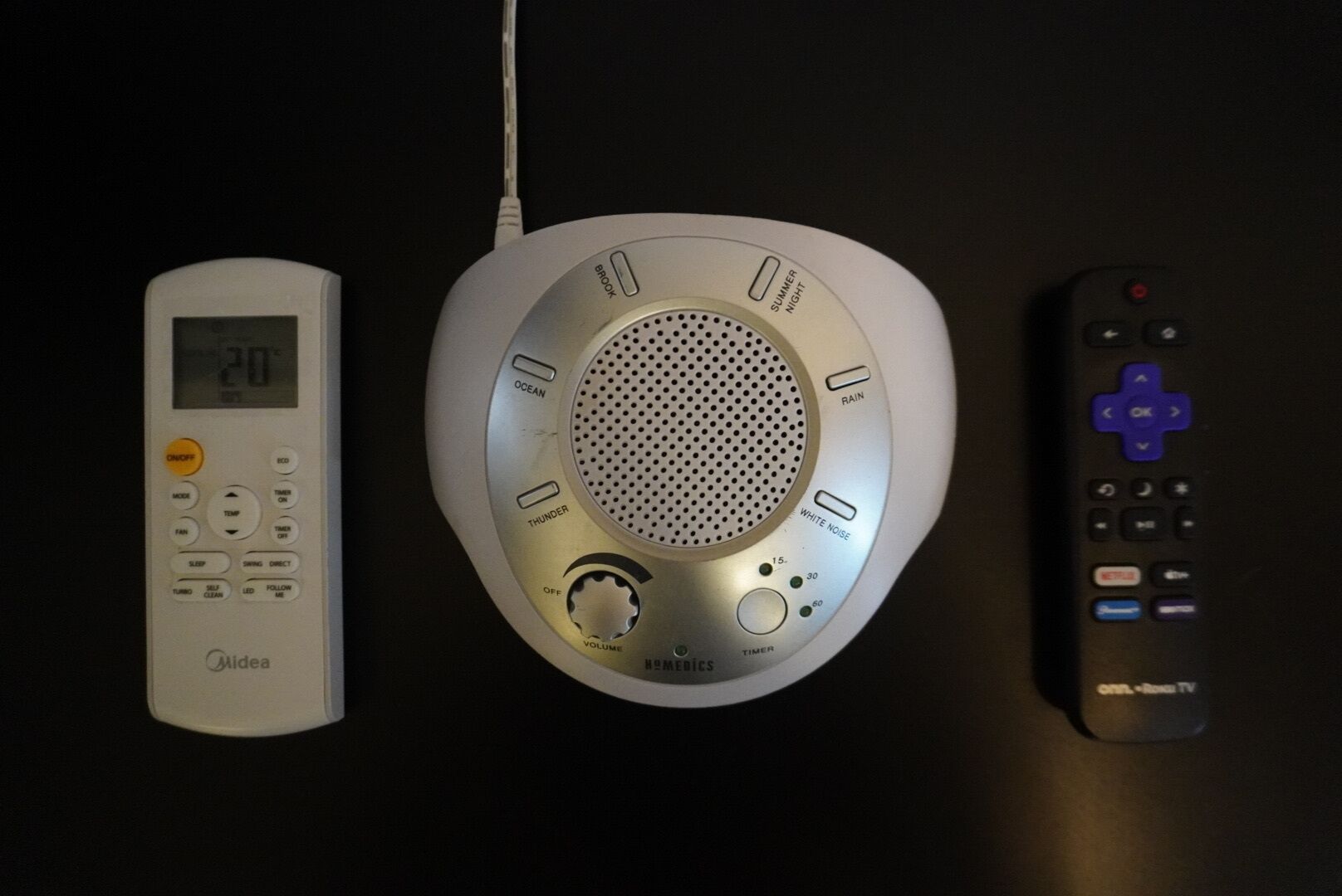 A white bedside table with a digital clock, a white bedside speaker, and a black remote control.