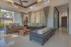 A modern living room with a sofa, ottoman, and coffee table. There is a statue on a small table, and a large window with curtains. - Neo Retro (Lonavala)