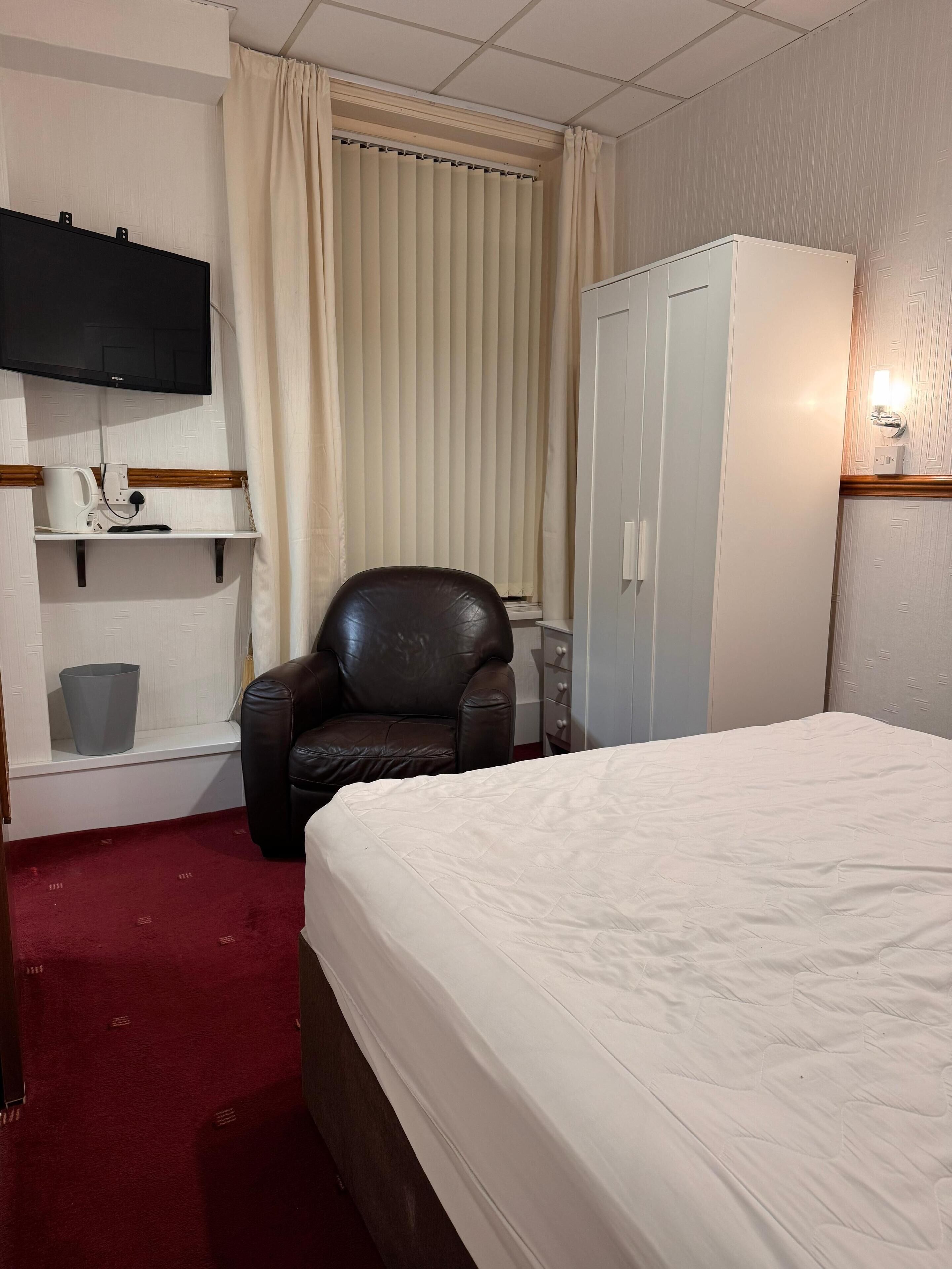 A hotel room with a bed, a leather chair, a wardrobe, a television, and a shelf with a kettle and a trash bin.