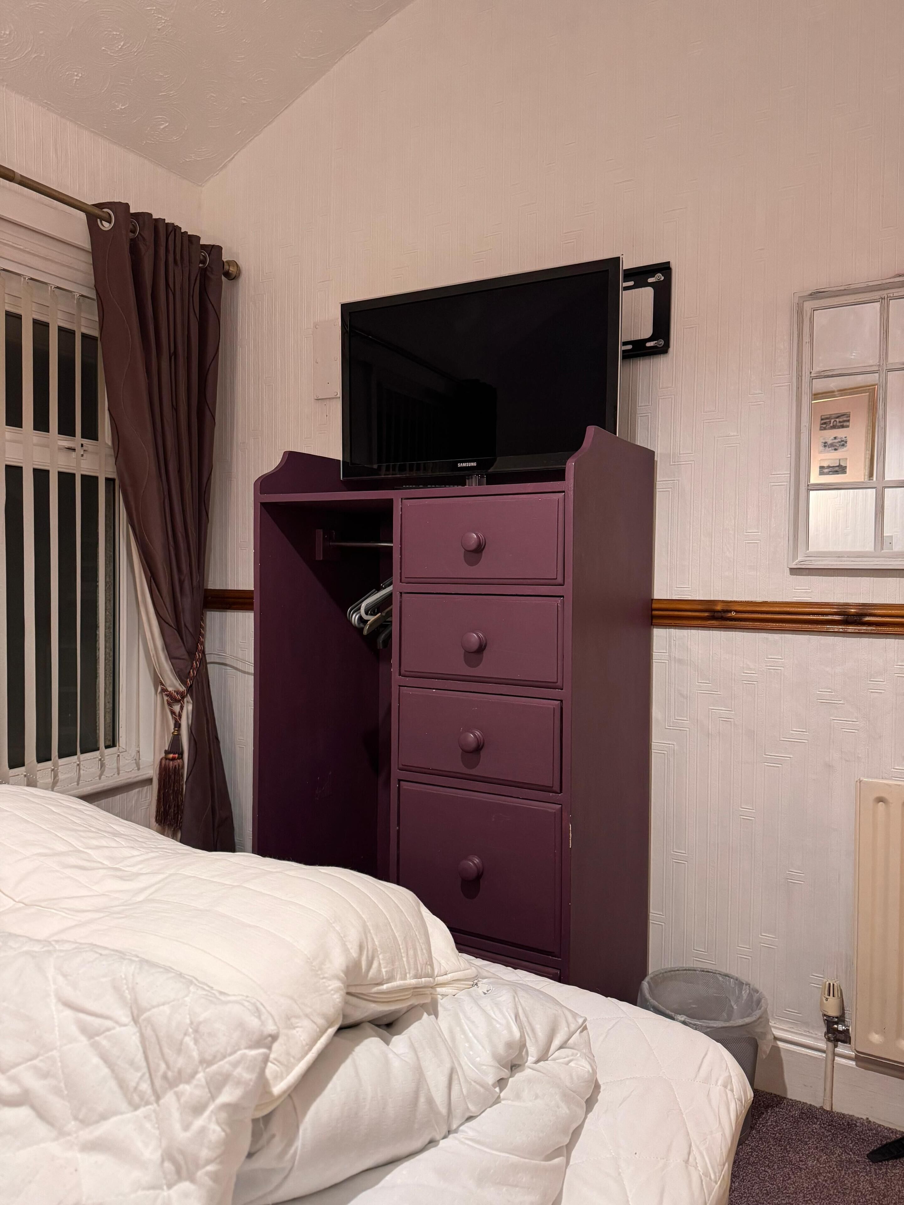 Una camera da letto con un letto, una cassettiera con cassetti, un televisore sopra e una finestra con tende, oltre a un termosifone.
