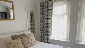 A neatly made bed with a brown blanket, a white duvet, and two pillows with a duck pattern. There is a small bedside table with a lamp, a window with striped curtains, and a mirror on the wall. - No 34 Vance Road (Blackpool)
