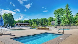 A rectangular pool with a stone border, surrounded by lounge chairs and umbrellas, with a covered seating area and trees in the background.