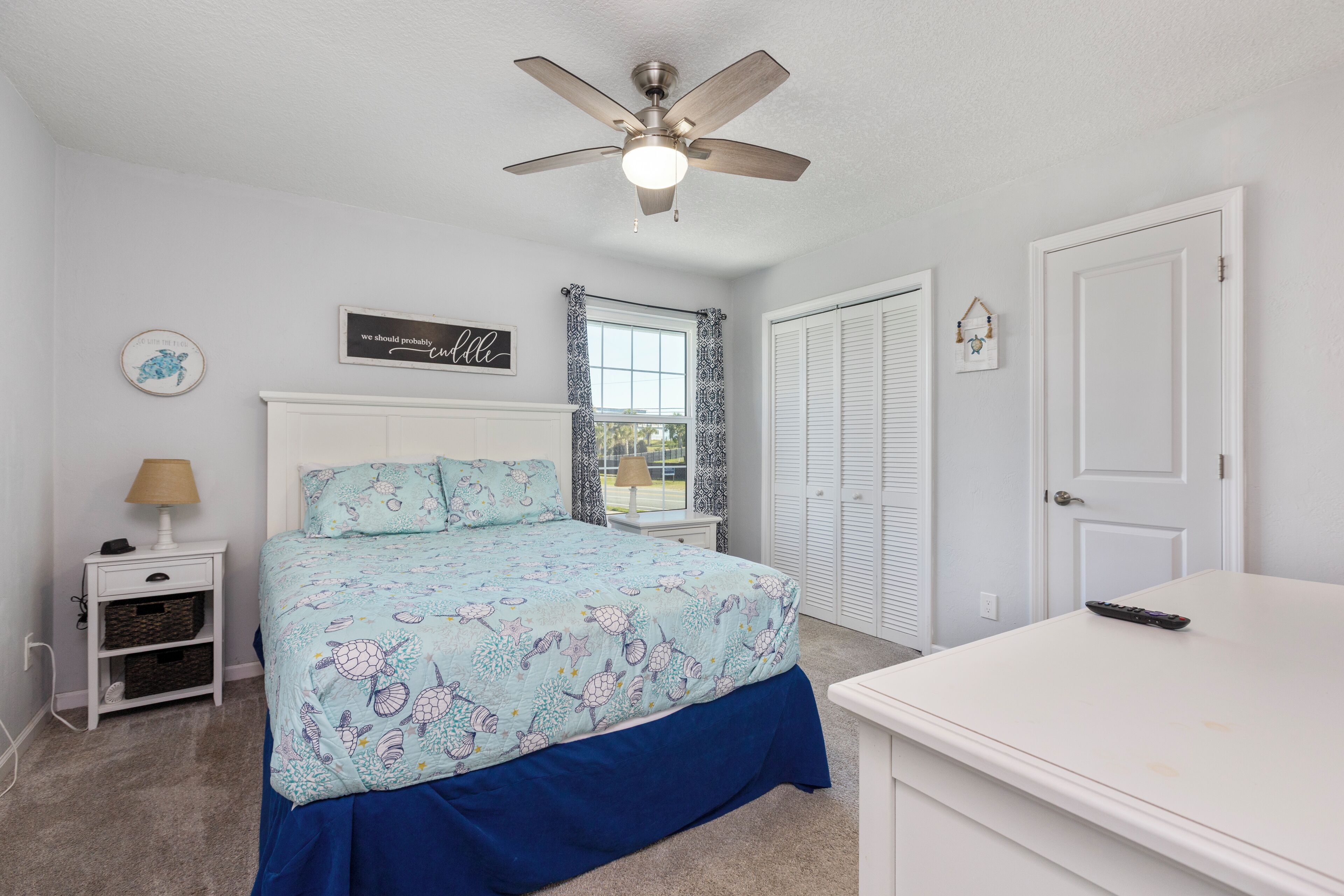 A bedroom with a bed, a ceiling fan, a nightstand, a dresser, and a window with curtains.