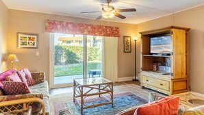 A living room with a ceiling fan, a glass coffee table, a sofa with patterned cushions, a wooden entertainment center, and a view of a garden through a sliding glass door. - Paws Welcome at Ponce Landing 25 (St. Augustine)
