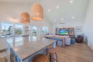 A spacious living area with a large island, wicker pendant lights, a flat-screen TV, and a view of the outdoors.