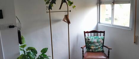 A room with a wooden floor, a chair with a floral cushion, a floor lamp with a hanging plant, and a window with blinds.