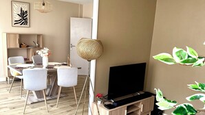 Ein modernes Wohnzimmer mit Holzboden, einem Flachbildfernseher auf einem Holzbodenständer, einem Essbereich mit weißem Tisch und Stühlen sowie einer großen gewebten Stehlampe.