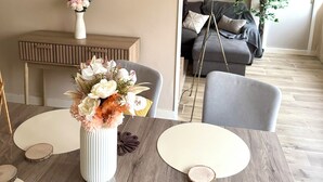 Ein Essbereich mit einem Holztisch, einer weißen Vase mit Blumen und einer grauen Couch im Hintergrund.