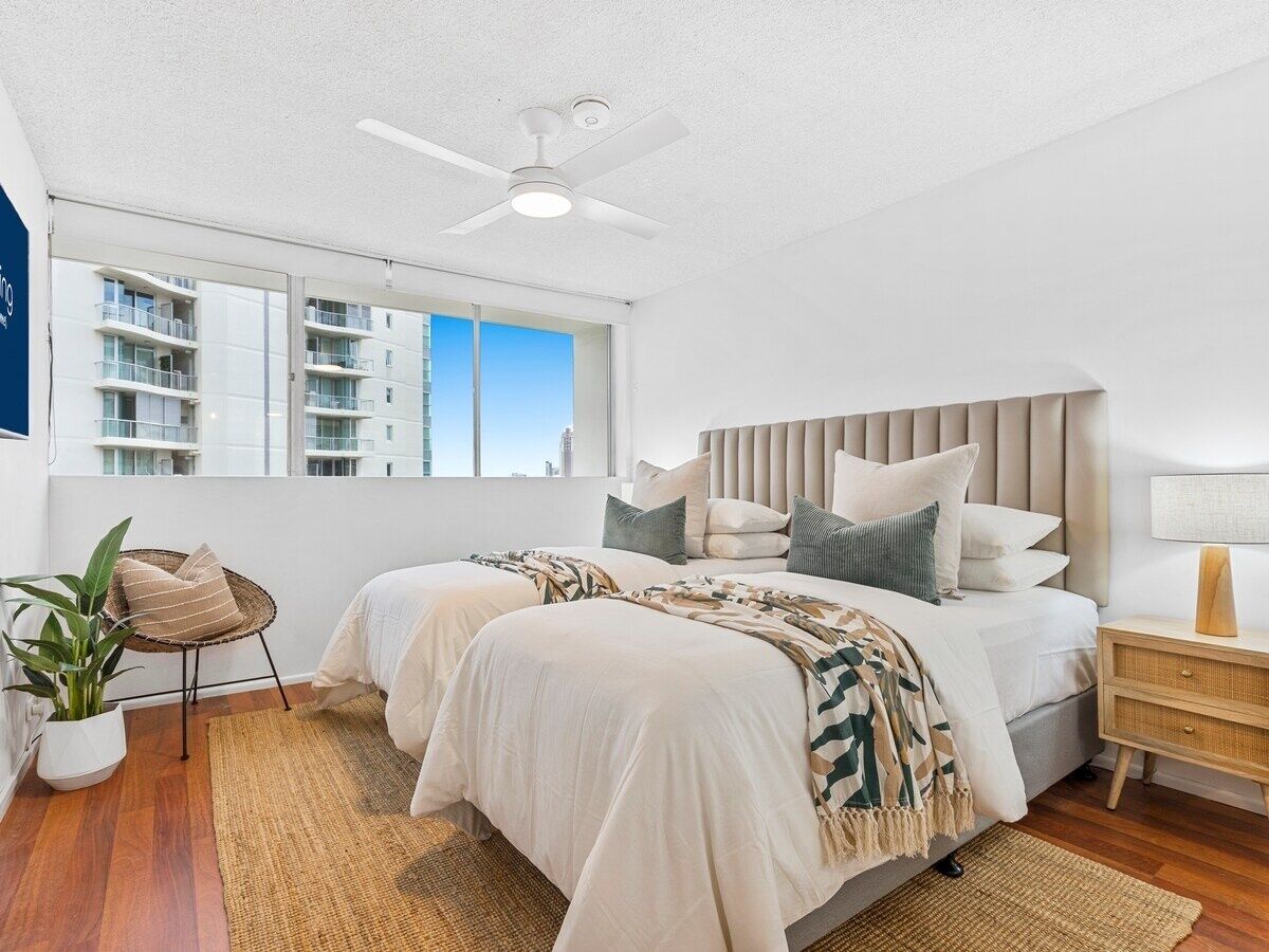 A modern bedroom with two beds, a ceiling fan, a large window, and a view of buildings.