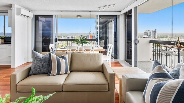 A modern living room with a beige sofa, a dining table with chairs, and a balcony with a view of the city. - 12th Floor Riverfront Retreat Views Walk to Beach (Surfers Paradise)