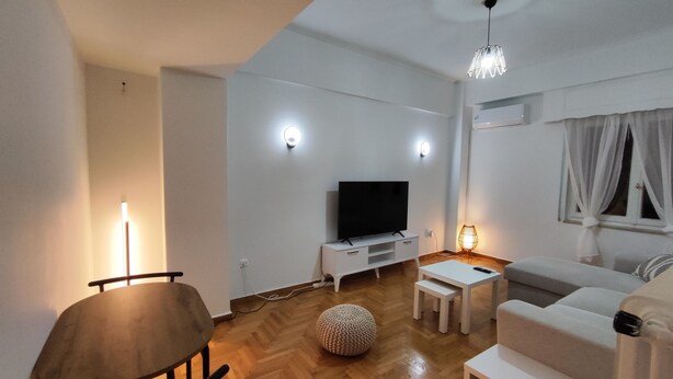 Een moderne woonkamer met een houten vloer, een witte bank, een televisie op een witte dressoir, een eettafel met stoelen, en een lamp.