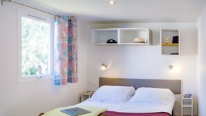 A neatly made bed with a laptop and folded clothes, a window with curtains, and built-in shelves with items. - Waterfront mobile home (Saint-Florent)