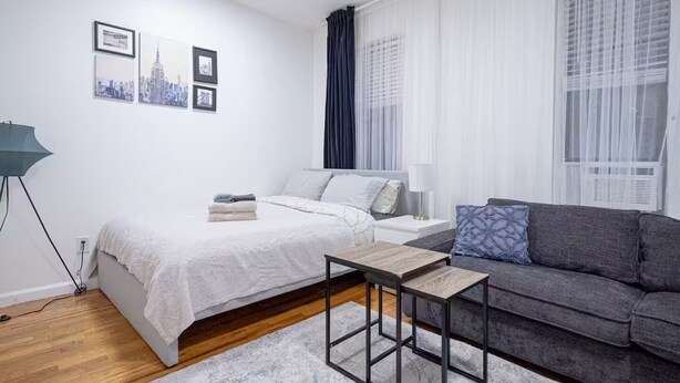 Un dormitorio moderno y bien iluminado con cama, sofá gris, una mesita y un cuadro con vista a la ciudad.