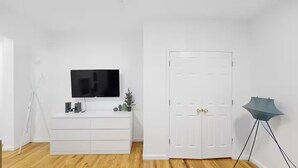Una habitación con un armario blanco, un televisor de pantalla plana sobre un aparador blanco, una puerta blanca y una lámpara de pie.