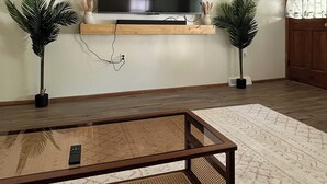 A living room with a flat-screen TV mounted on the wall, a wooden shelf below, and two potted plants on either side. - Welcome to the Butterscotch Bungalow! (Hummelstown)