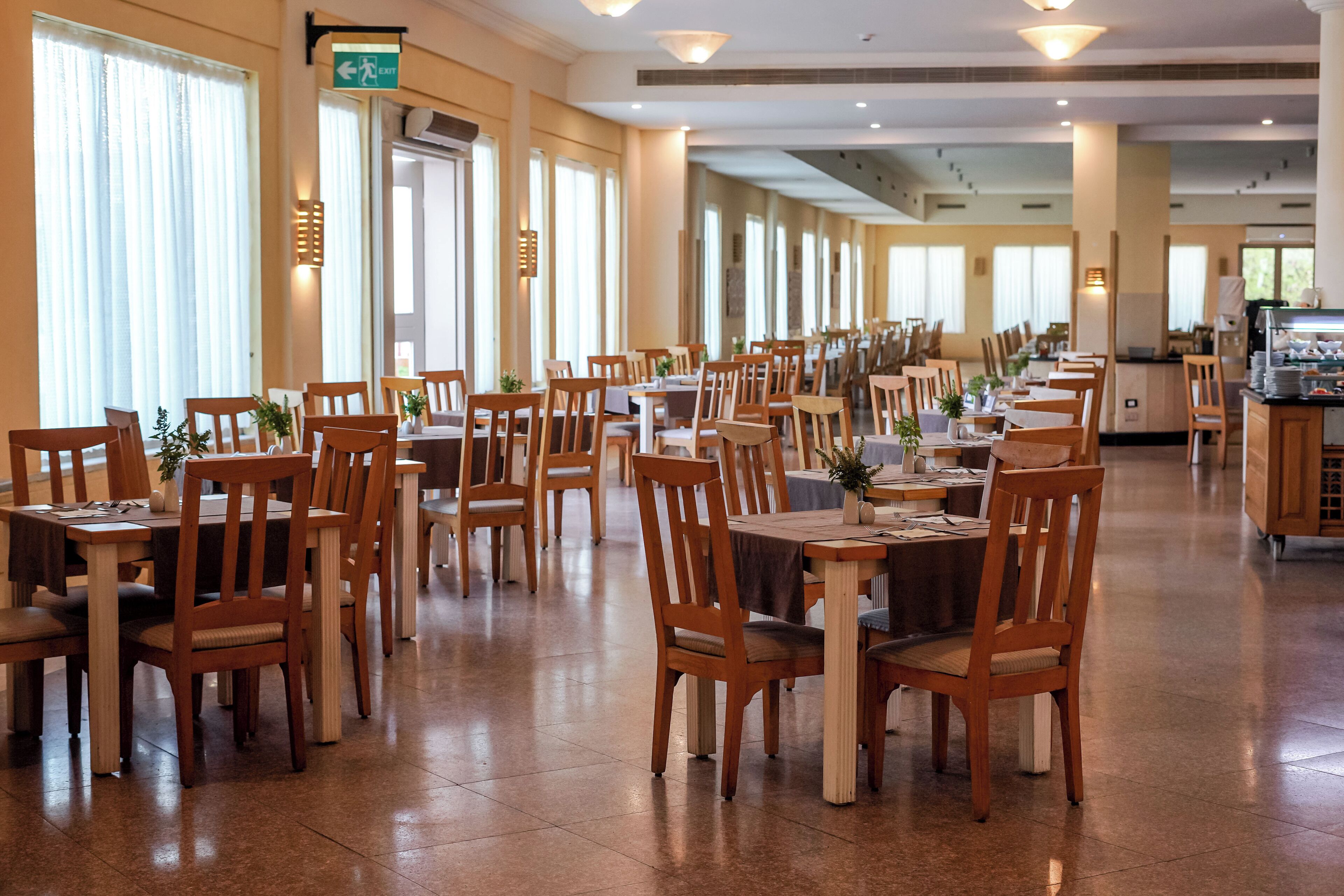 Un comedor amplio con varias mesas y sillas, ventanas grandes y una señal de salida.