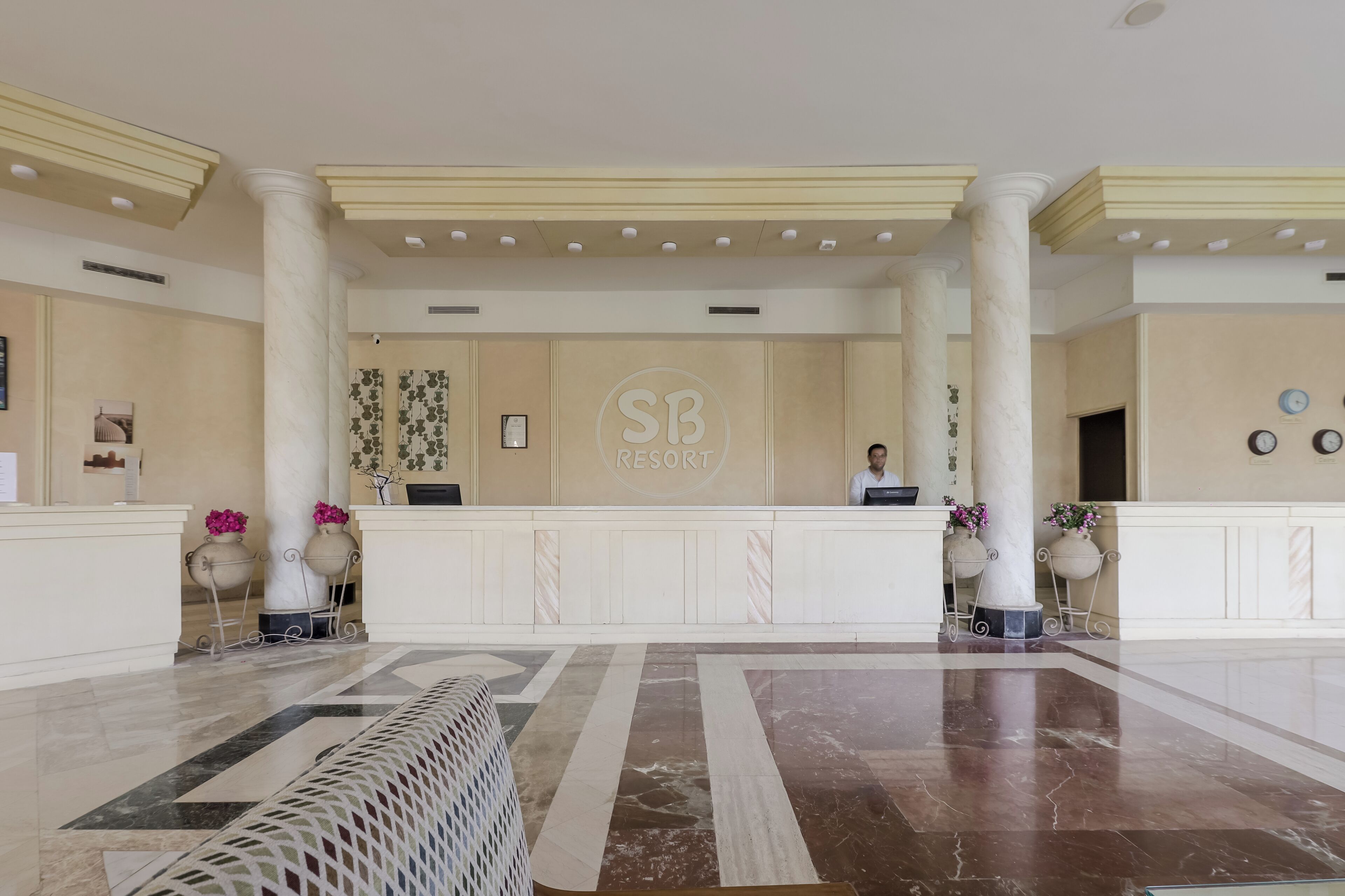 A hotel reception area with a marble floor, a reception desk, and a large 'SB' logo on the wall.
