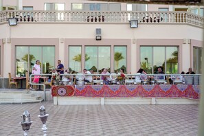 An outdoor dining area with tables and chairs, a decorative railing, and a building with large windows in the background. - Shoni Bay Resort (Marsa Alam)