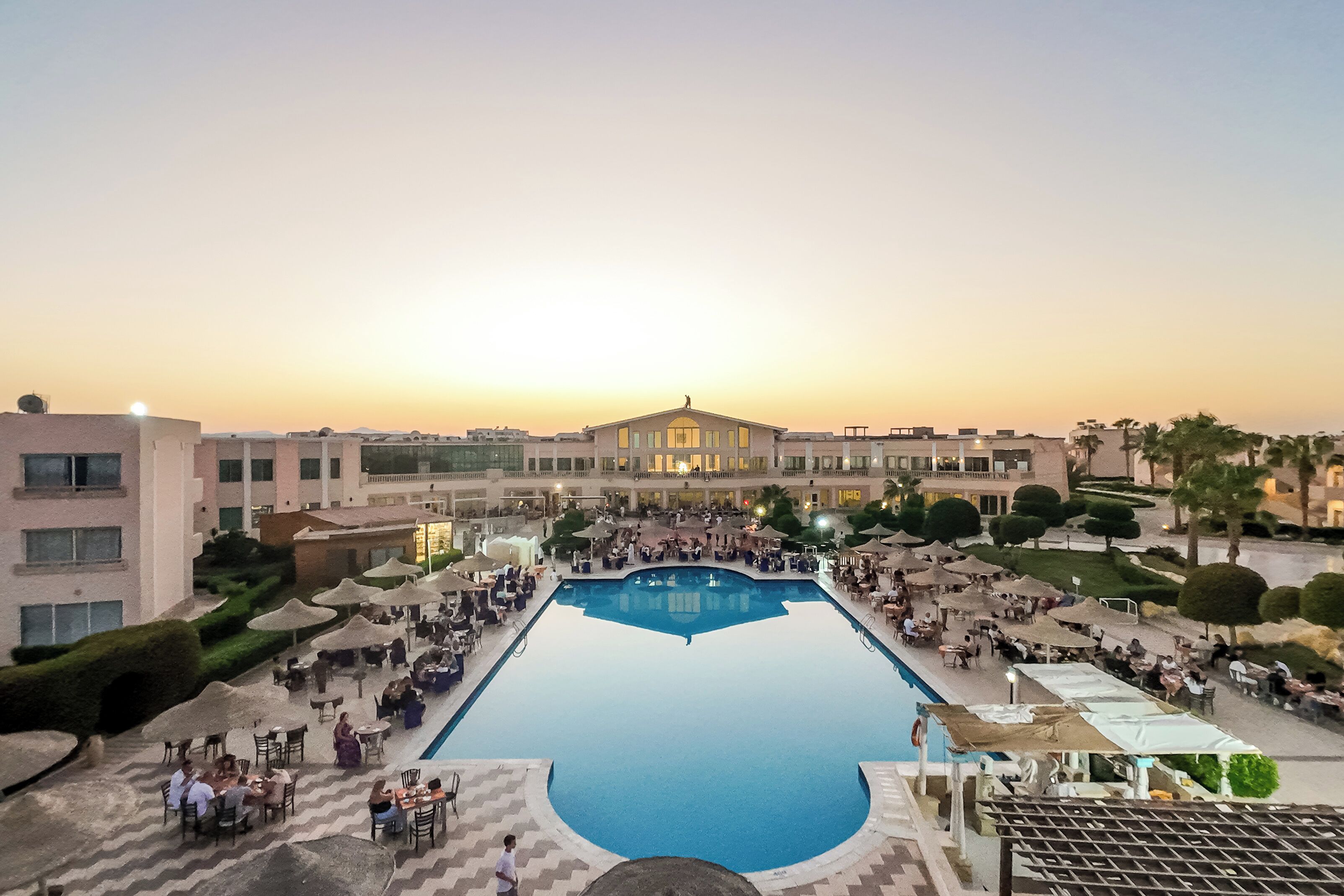 A resort with a central swimming pool surrounded by outdoor seating areas and a large building in the background.