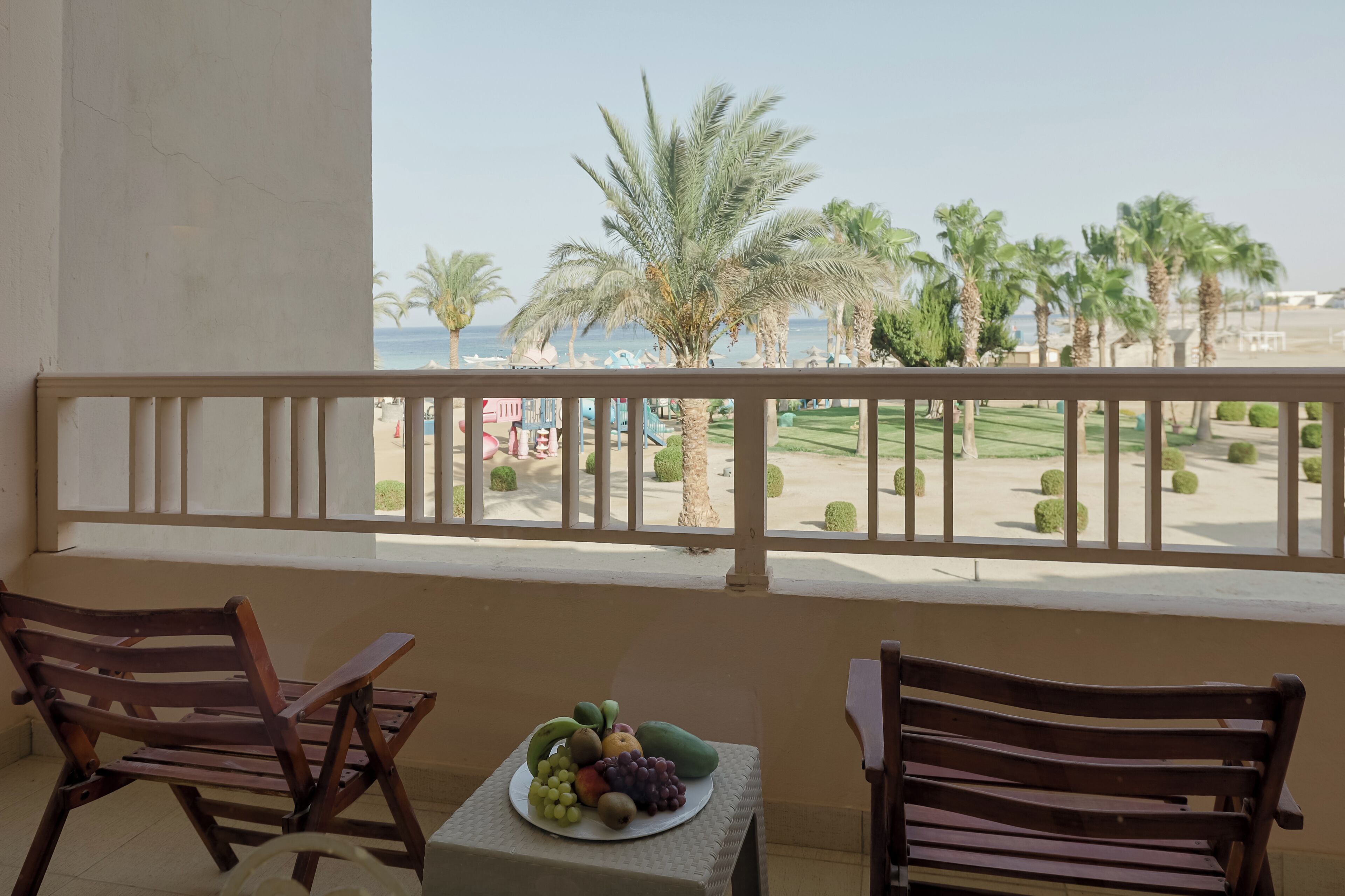A balcony with two wooden chairs, a small table with a fruit plate, and a view of a beach with palm trees and a clear sky.