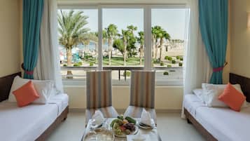 Een hotelkamer met twee bedden, een eettafel met fruit en glazen, en uitzicht op een strand en palmbomen.