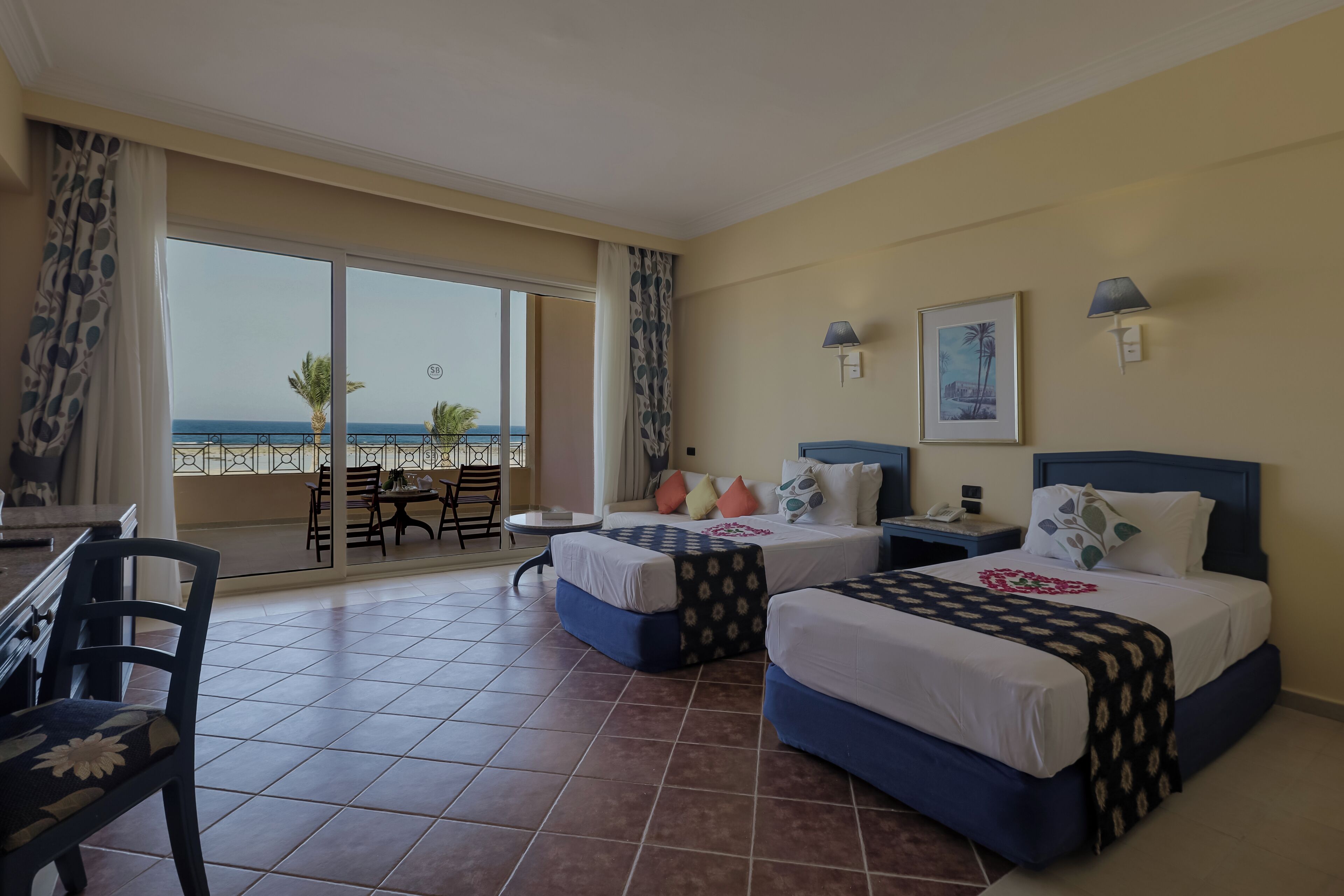 A hotel room with two beds, a balcony with a view of the sea, and a dining area with chairs and a table.