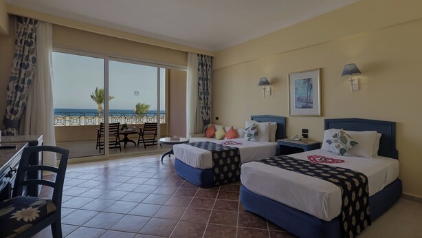 Quarto de hotel com duas camas, varanda com vista para o mar e área de refeições com cadeiras e mesa.