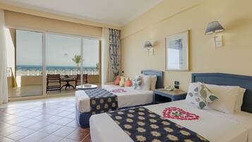 A hotel room with two beds, a balcony with a view of the sea, a dining area with chairs and a table, and a large window.