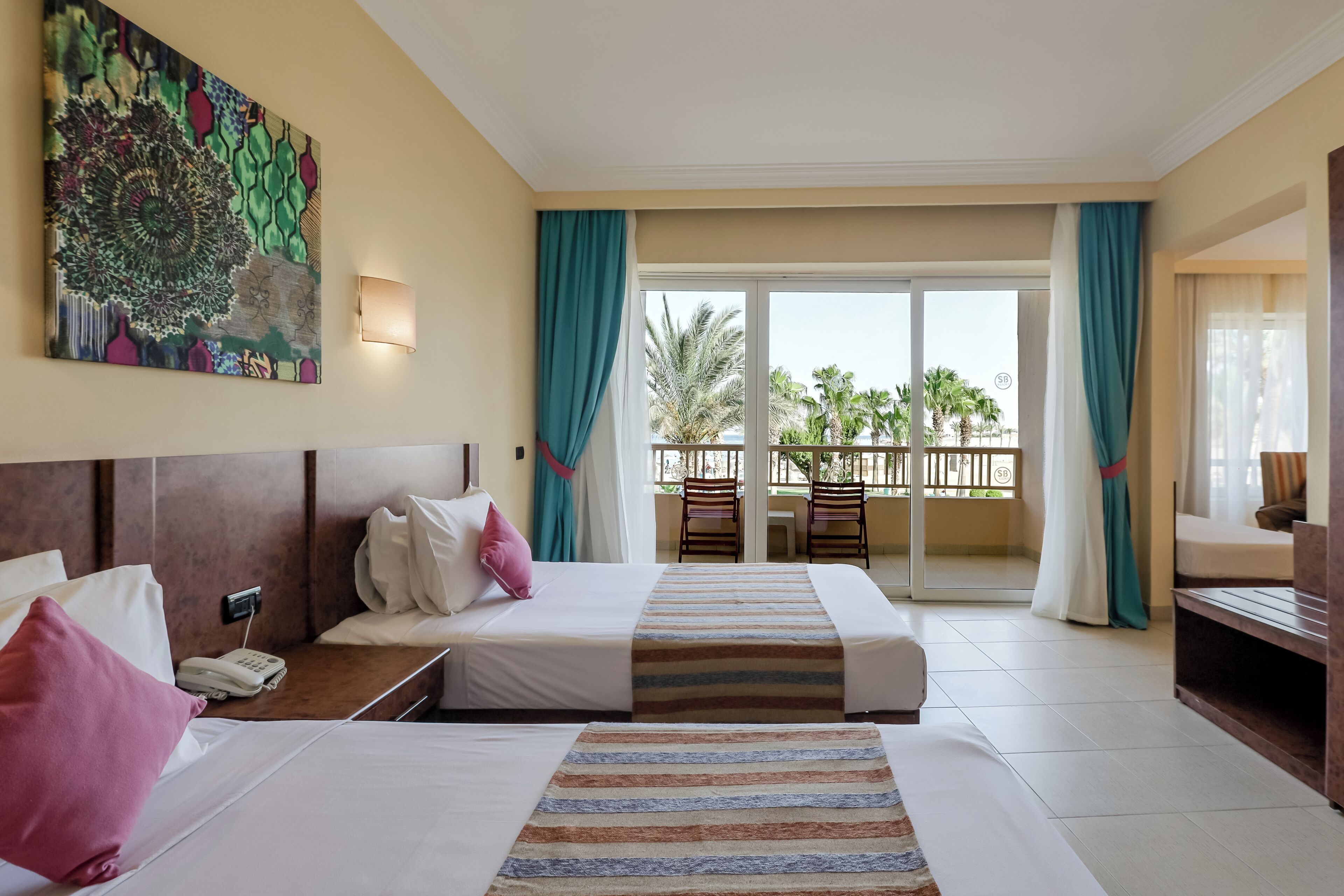 A hotel room with two beds, a painting on the wall, a balcony with chairs, and a view of palm trees.
