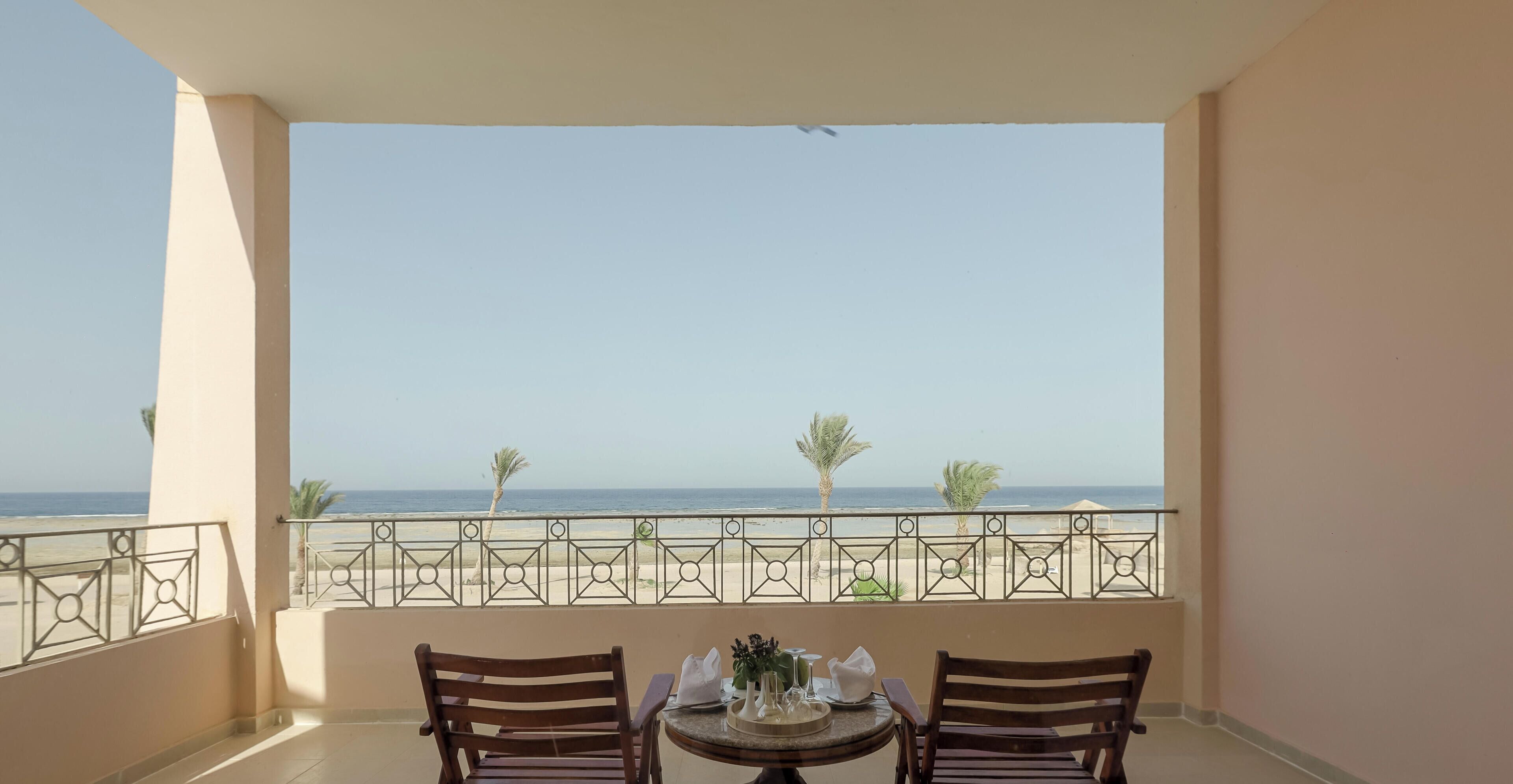 A balcony with a table and chairs, overlooking a beach with palm trees and the sea.