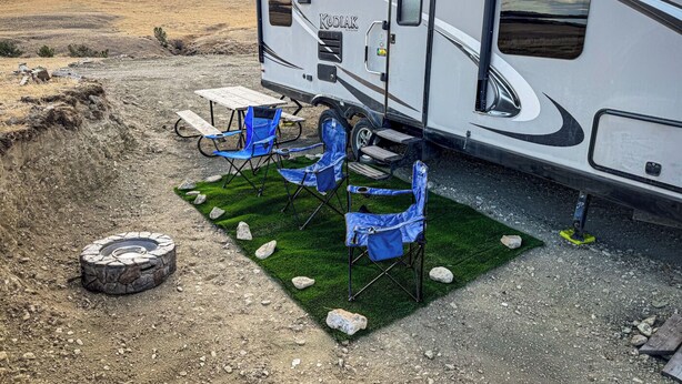 A recreational vehicle (RV) with a picnic table and blue folding chairs set up on a grassy patch beside a dirt road. - Crazy Mountain Camper NE of Livingston (Livingston)