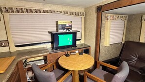A compact room with a wooden table and two chairs, a television on a stand, and a window with blinds. - Crazy Mountain Camper NE of Livingston (Livingston)