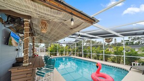 An indoor swimming pool with a wooden bar and chairs, surrounded by a covered area with a view of greenery.