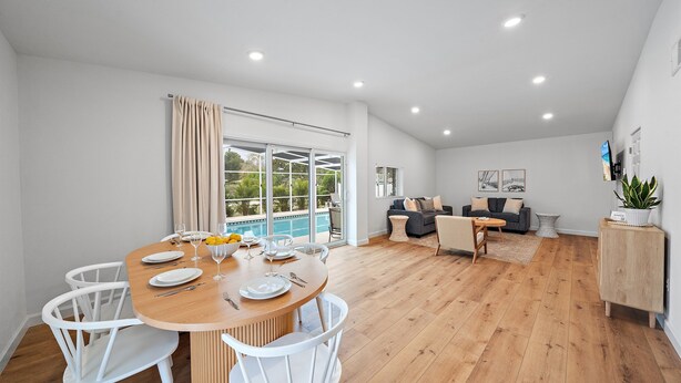 A spacious living area with a wooden dining table set for four, white chairs, a grey sofa, and a small side table.