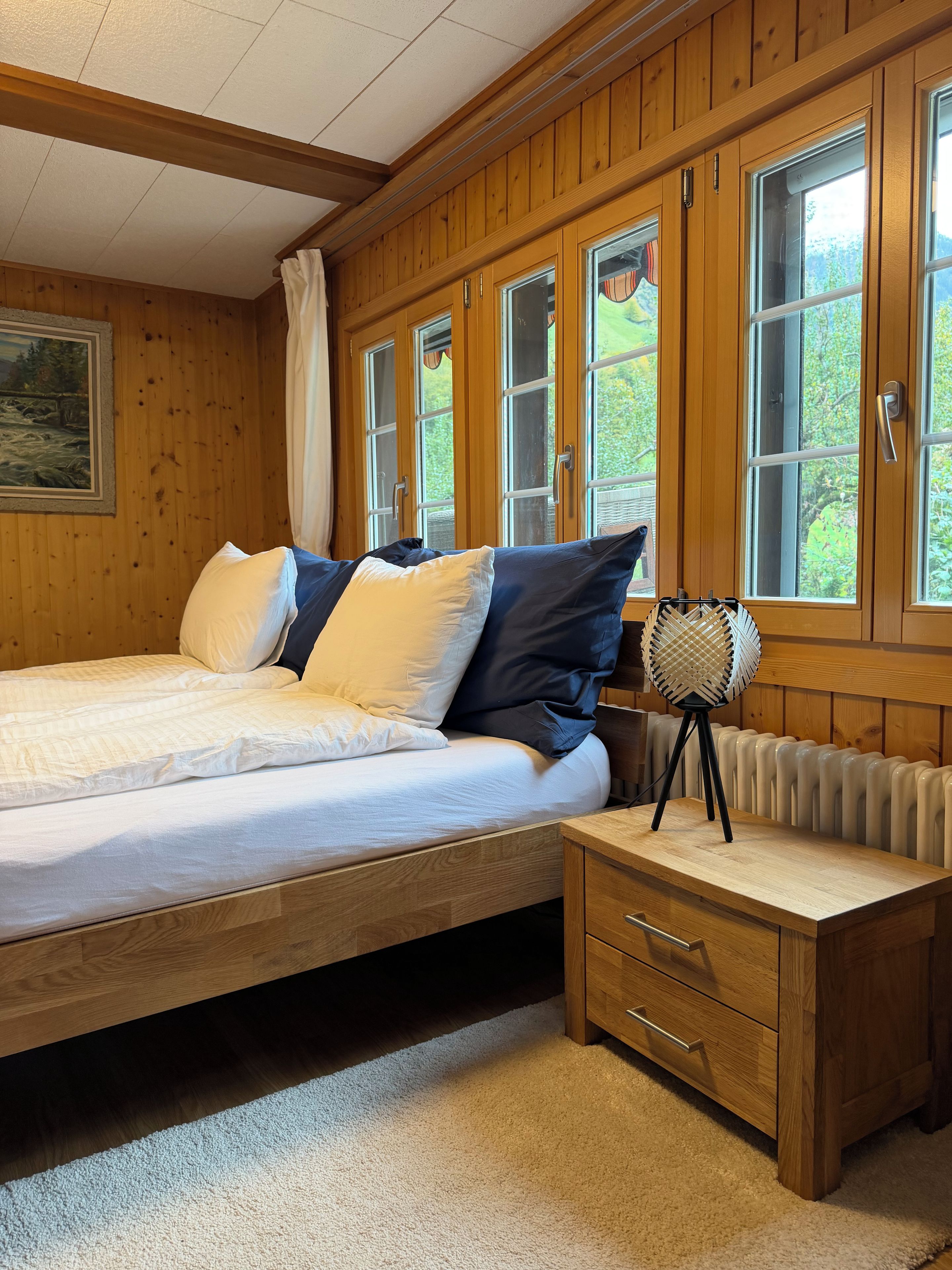A wooden-paneled bedroom with a bed, a bedside table, a radiator, and a window with curtains.