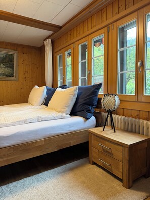 A wooden-paneled bedroom with a bed, a bedside table, a radiator, and a window with curtains.