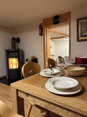 A dining area with a wooden table set for two, a wood-burning stove, and a mirror reflecting the room.