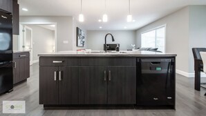 A modern kitchen with dark cabinetry, a built-in dishwasher, pendant lighting, and a large island. - Luxury Executive 4BD 2.5B AC home in the exclusive neighbourhood of Windermere (Edmonton)