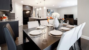 A modern kitchen with dark cabinetry, a central island, and a dining area with white chairs and a dark table. - Luxury Executive 4BD 2.5B AC home in the exclusive neighbourhood of Windermere (Edmonton)