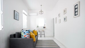 Een moderne woonkamer met een grijze bank, kleurrijke kussens, een gestreept vloerkleed en een eethoek met een witte tafel en stoelen.