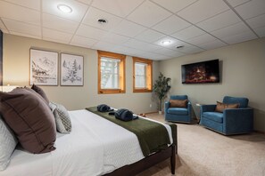 A hotel room with a bed, two blue armchairs, a large painting, and two windows with blinds.