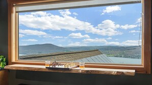 Un tablero de ajedrez y un vaso de líquido ámbar sobre un alféizar de madera, con vistas a un paisaje montañoso.